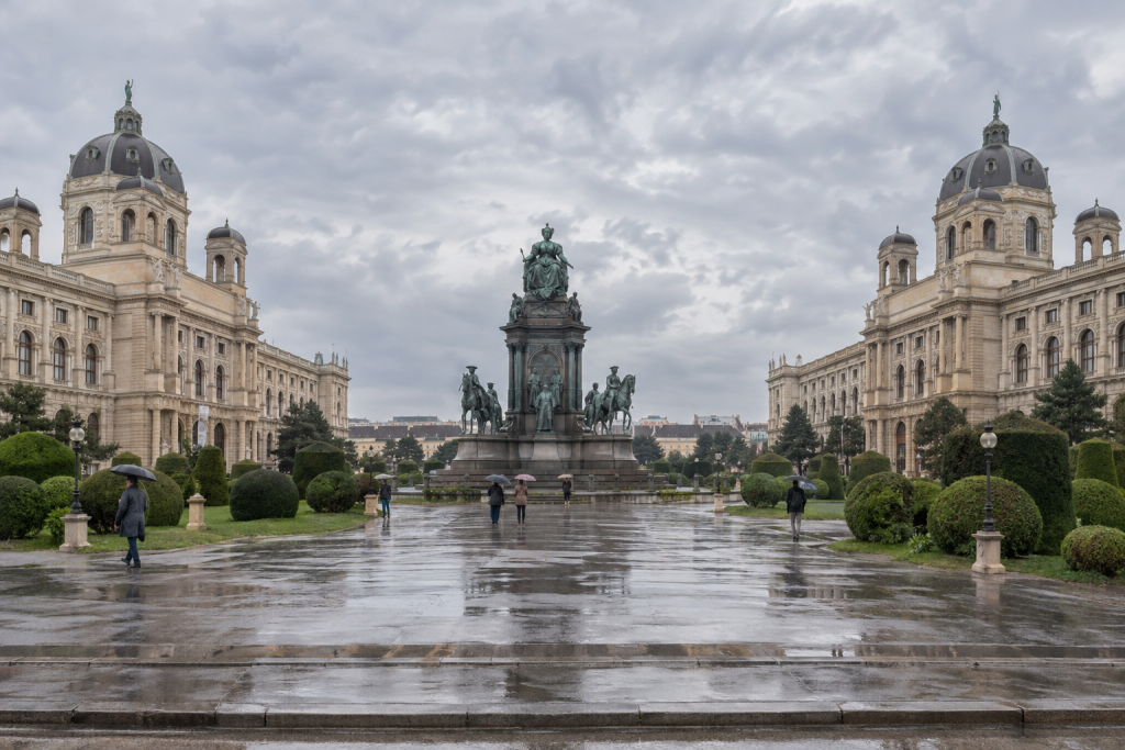 KI-generierte Ansicht des Maria-Theresien-Platzes mit zentralem Denkmal, zwei Museumsfassaden und Menschen mit Regenschirmen.