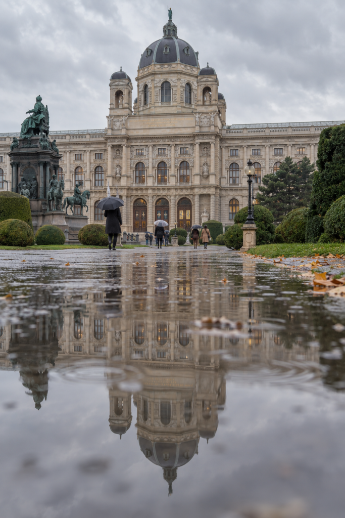 KI-generierte Museumsansicht mit Kuppel, die sich auf einem regennassen Weg am Maria-Theresien-Platz spiegelt.