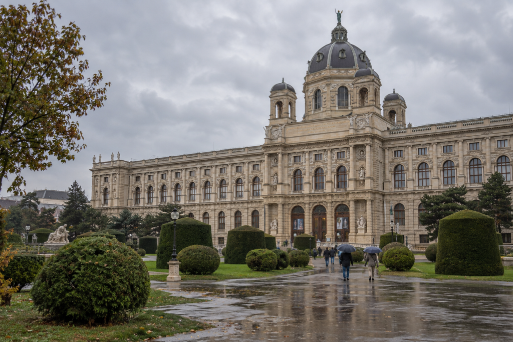 KI-generierte Museumsansicht mit grosser Kuppel, Gartenflächen und Menschen mit Regenschirmen auf nassen Wegen.