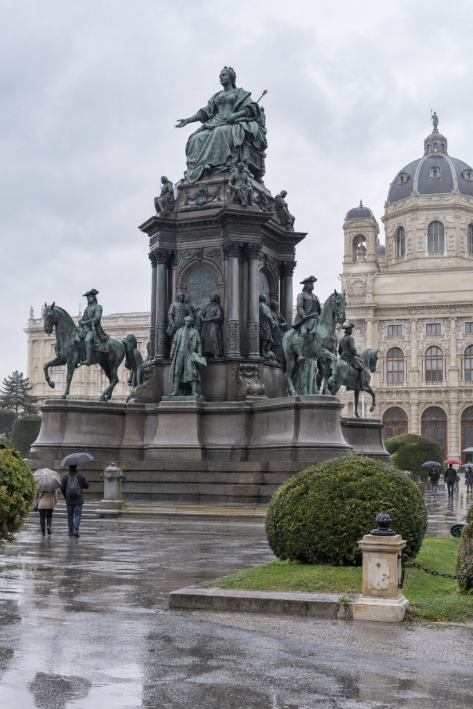 KI-generierte Denkmalansicht mit sitzender Herrscherin, hohem Sockel und Reiterfiguren auf einem regennassen Platz.