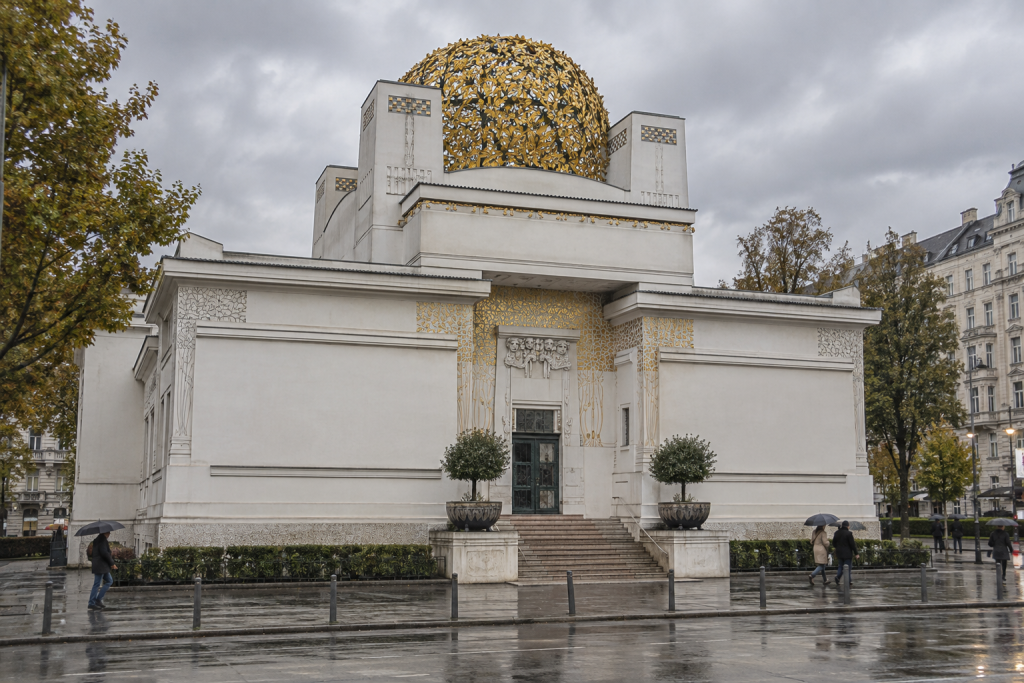 KI-generierte Ansicht der Secession mit hellen geometrischen Fassaden, goldener Blätterkuppel und nassem Vorplatz.