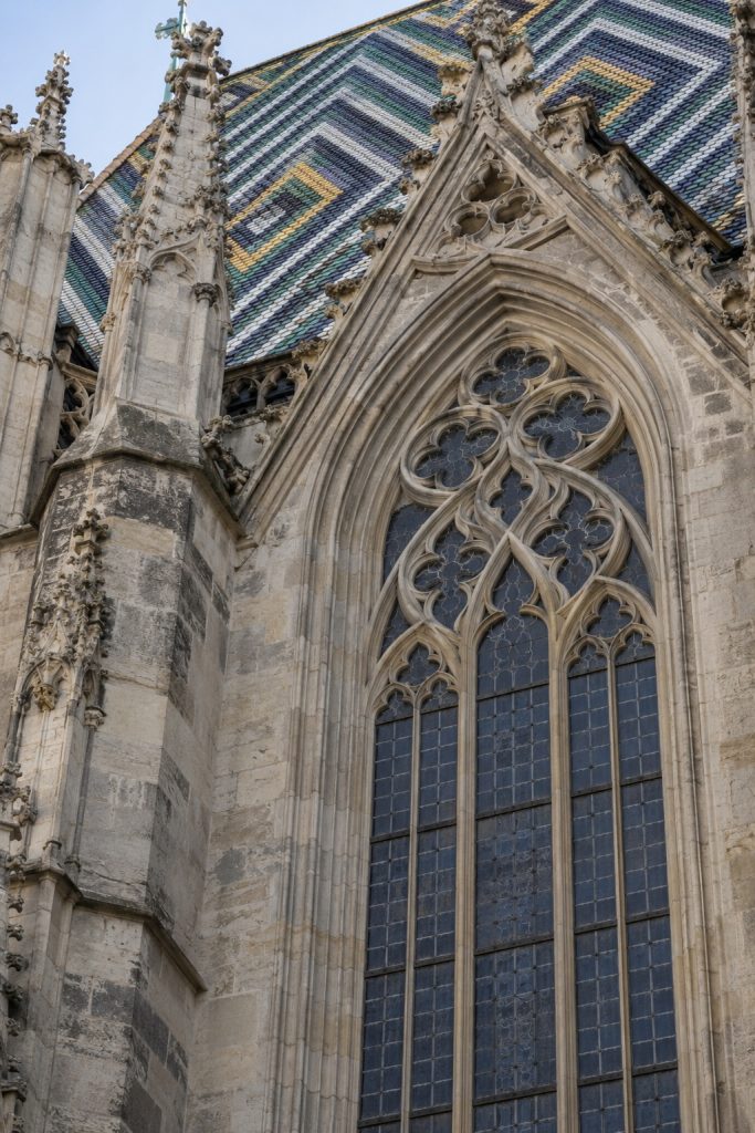 KI-generiertes Architekturdetail mit einem gotischen Masswerkfenster, verwittertem Stein und gemustertem Ziegeldach.